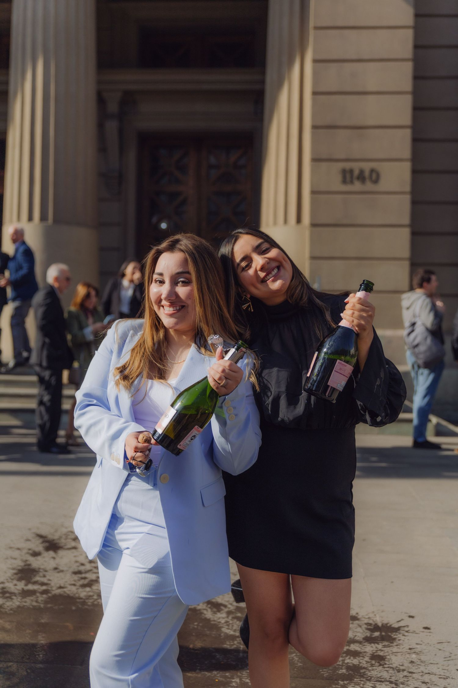Two young women celebrating with champagne bottles outside grand building entrance.