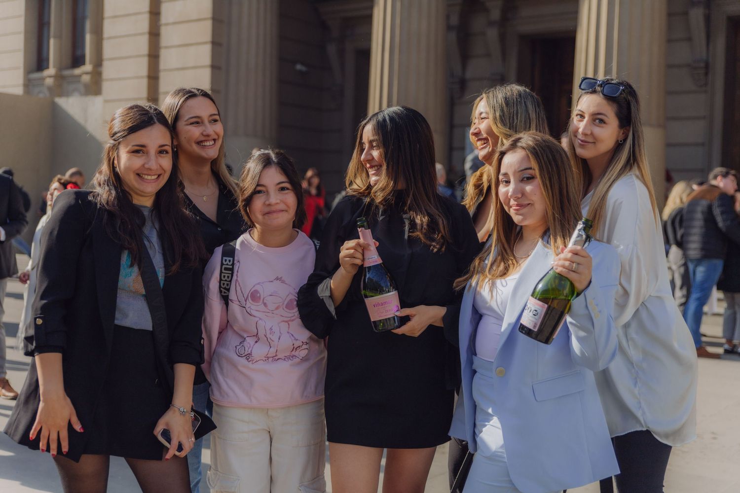 Seven young women posing together and smiling outside neoclassical building columns.
