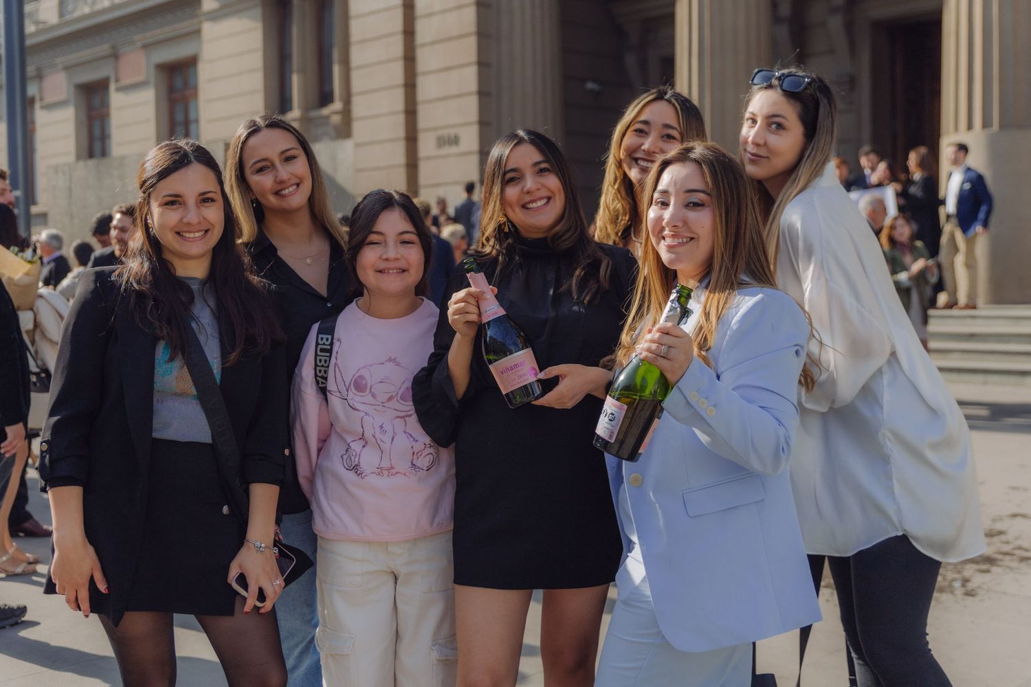 Group of seven young women smiling and celebrating with champagne outside building.