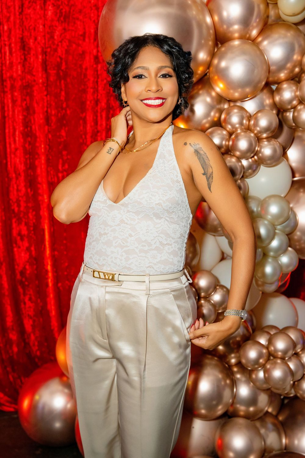 Woman in white lace top and cream pants posing confidently in front of gold balloons and red curtain backdrop.
