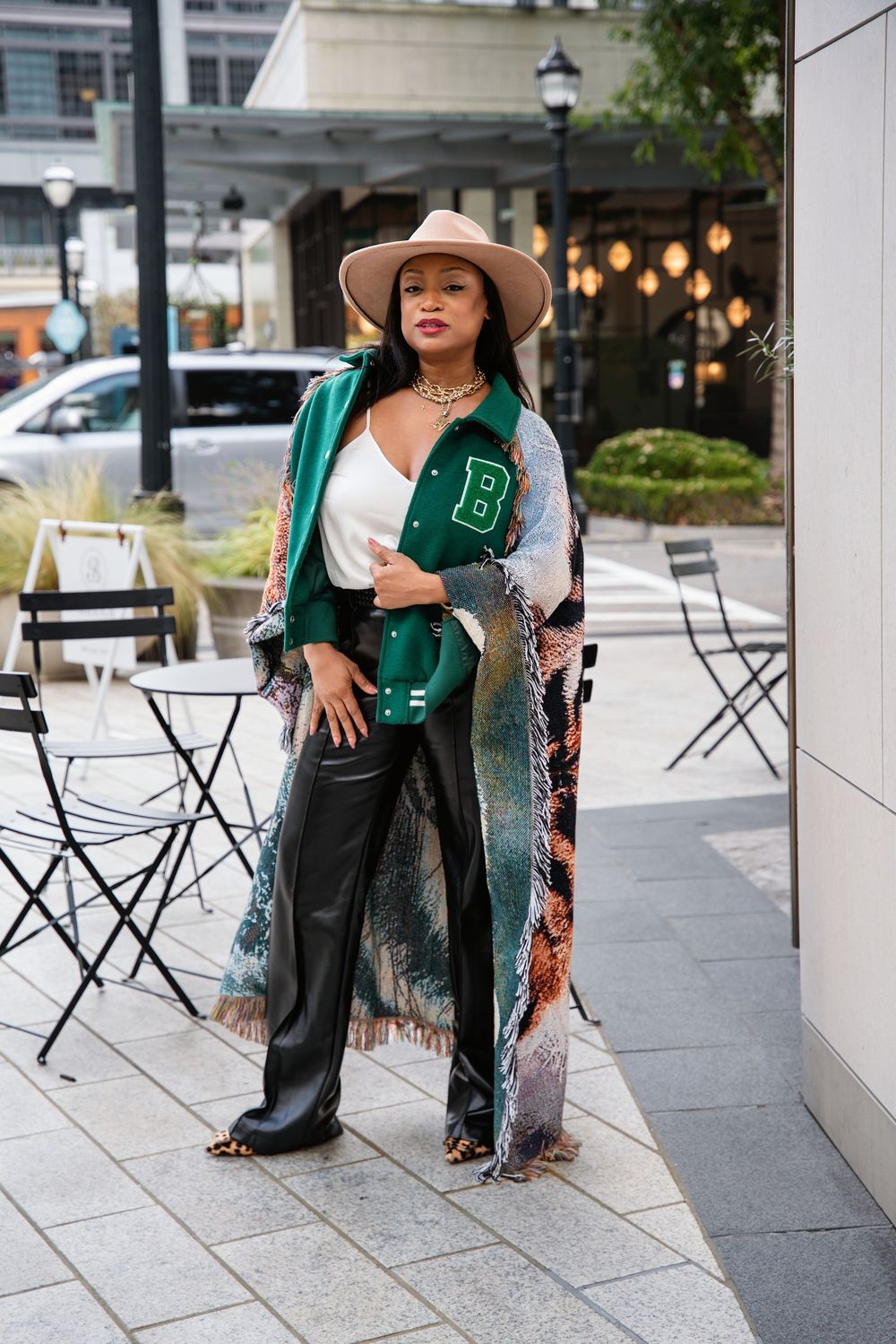 Woman in wide-brim hat, patterned kimono, leather pants, and leopard boots holding a green varsity bag outdoors.