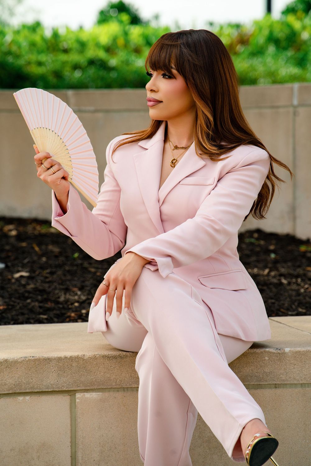 Woman in pink blazer suit holding a decorative hand fan, sitting outdoors with green foliage background.