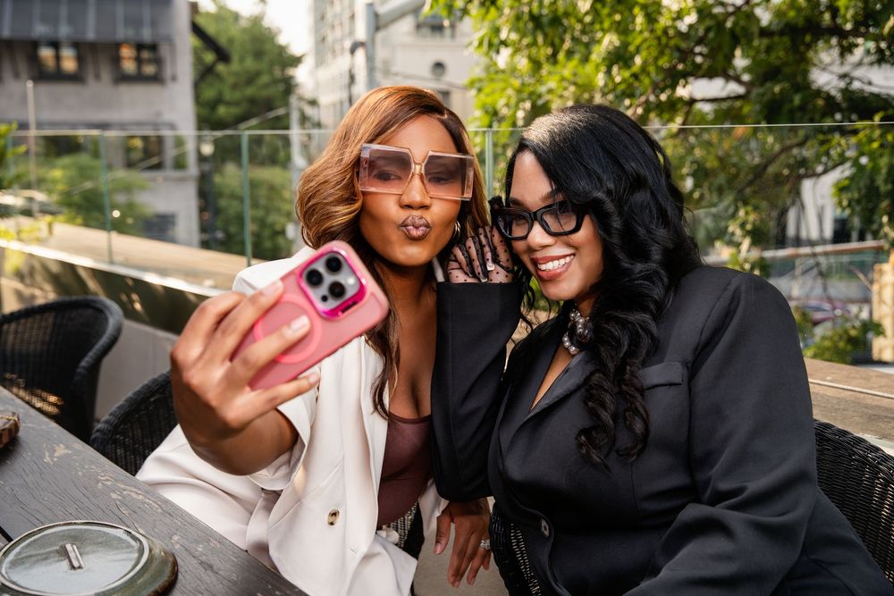Two stylish women in sunglasses take a selfie with a pink phone at an outdoor rooftop cafe.