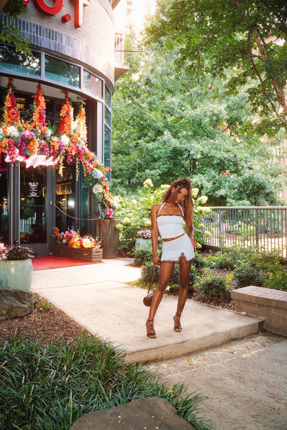 A woman in a white crop top and mini skirt walks outside a floral-decorated building surrounded by lush green trees.