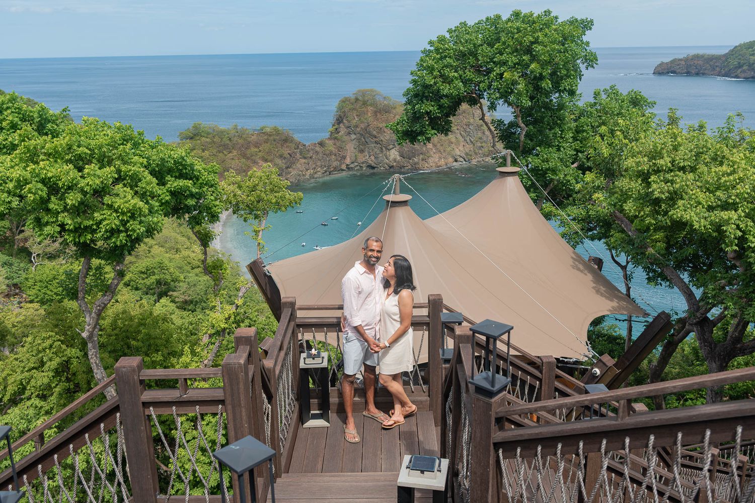 Couple smiling on wooden steps overlooking a tropical bay with lush greenery and a luxury tent resort below.
