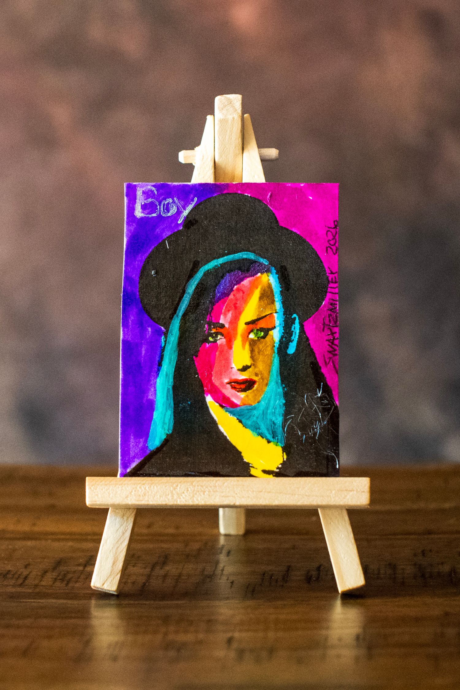 Colorful mini canvas painting of a woman in a black hat displayed on a small wooden easel.