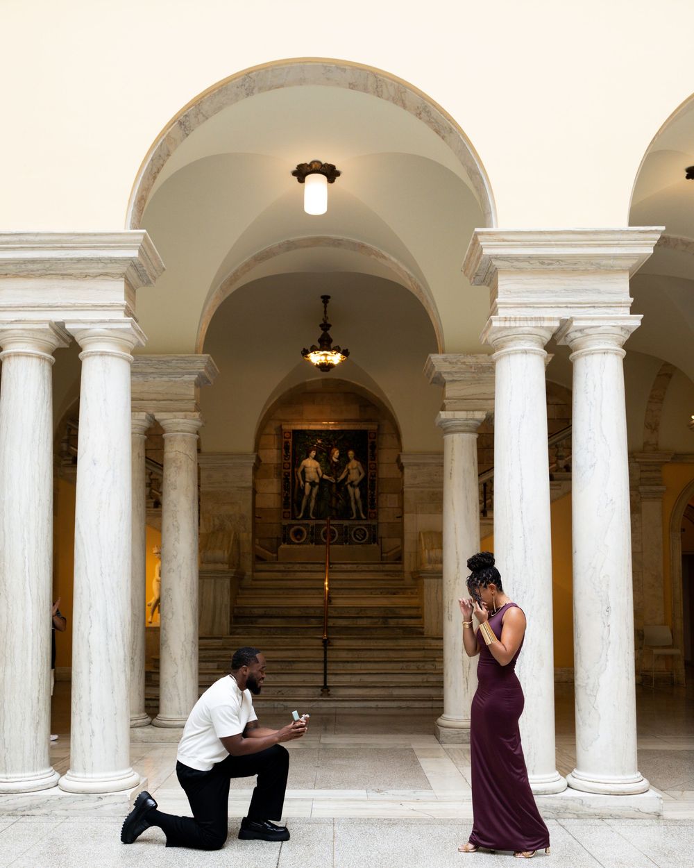 Man proposing on one knee to woman in burgundy gown inside elegant marble columned hall with arched ceilings.