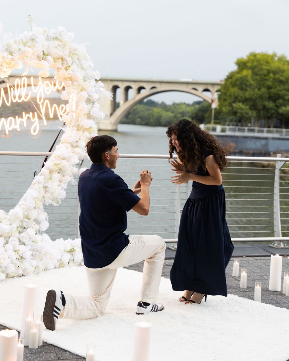 Man proposes on one knee to woman near waterfront with floral arch and Will You Marry Me neon sign.