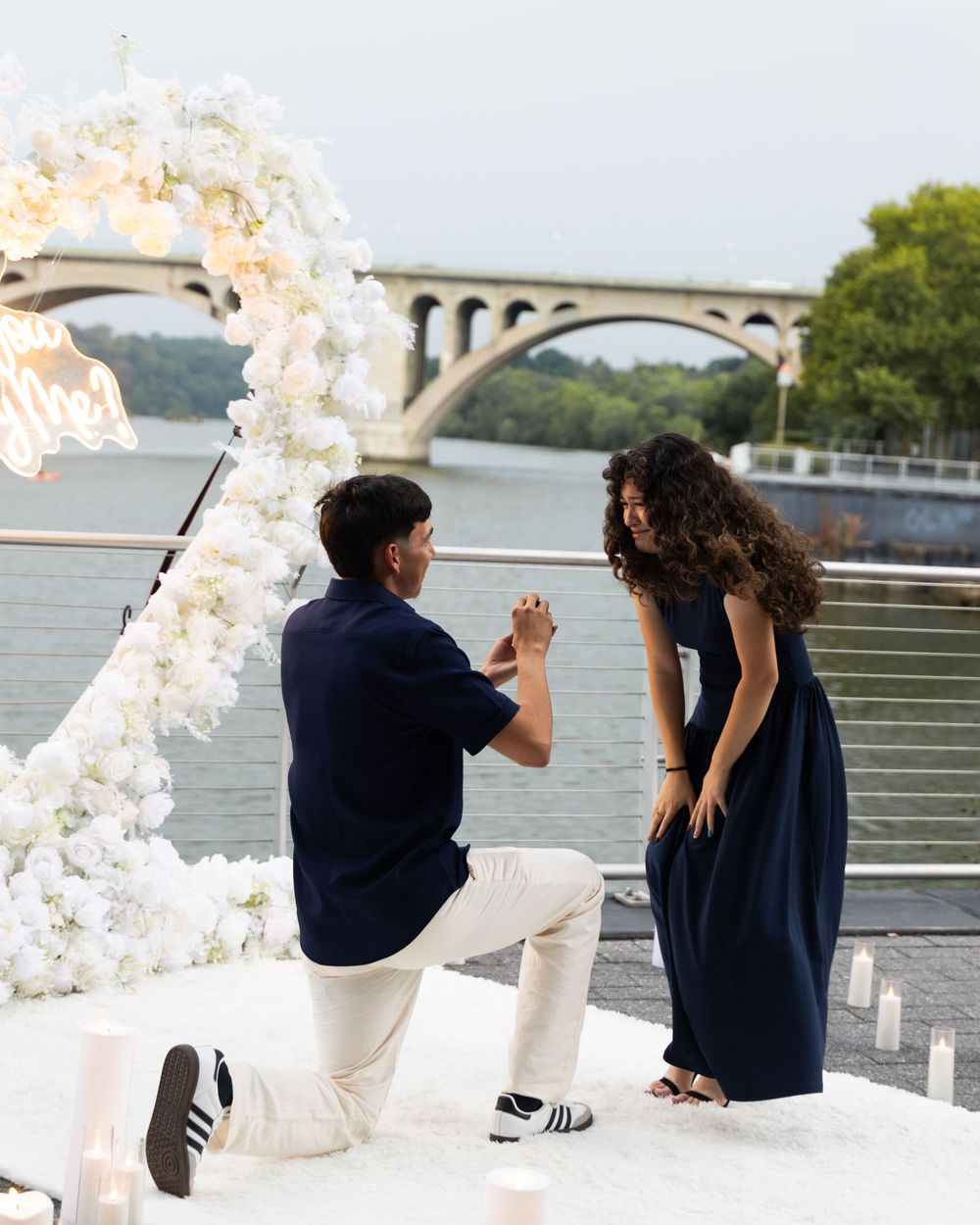 Man proposing to woman on one knee by a river with a floral arch and candles at sunset.