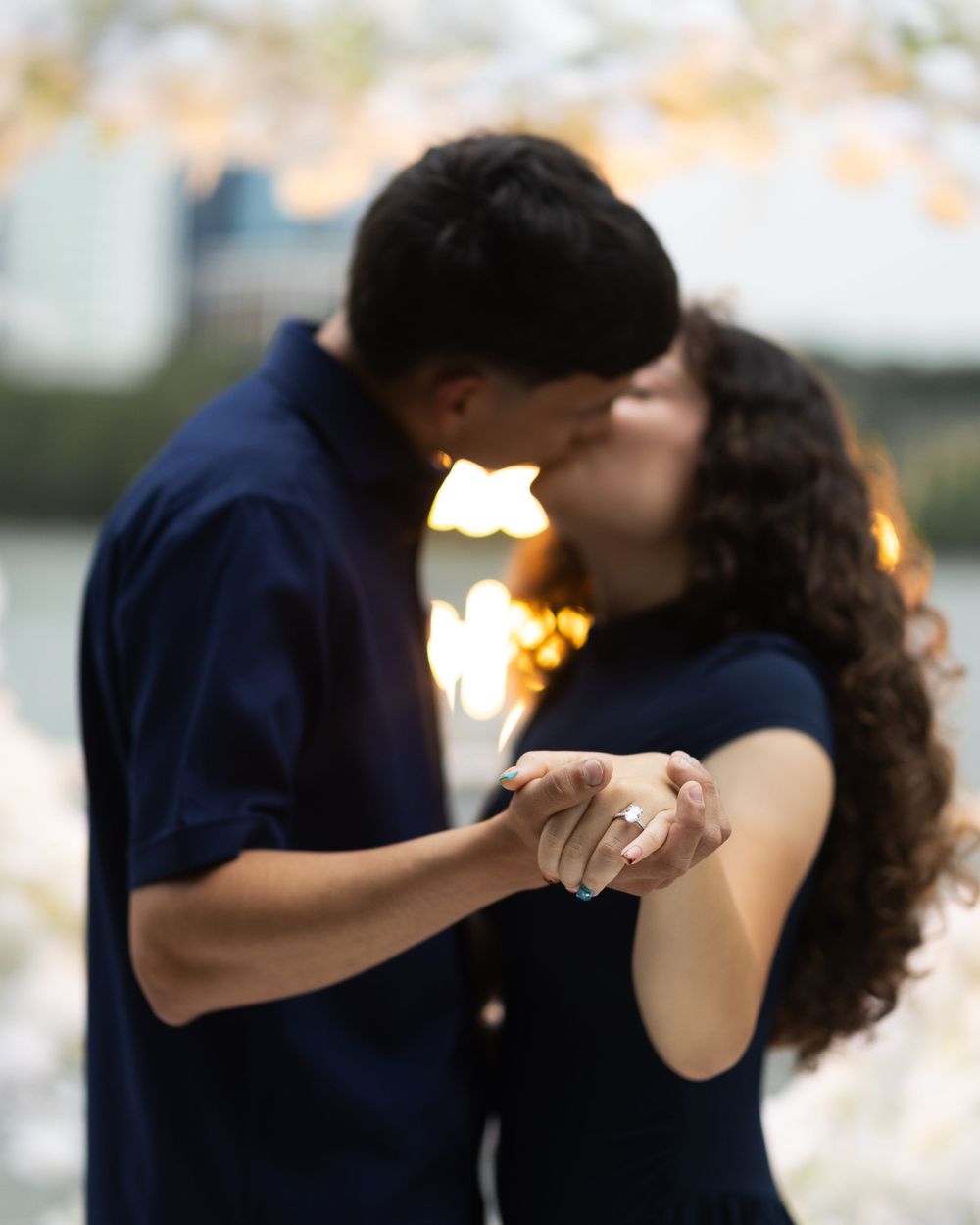 Couple sharing a romantic kiss while holding hands showing an engagement ring, with warm bokeh lights in the background.