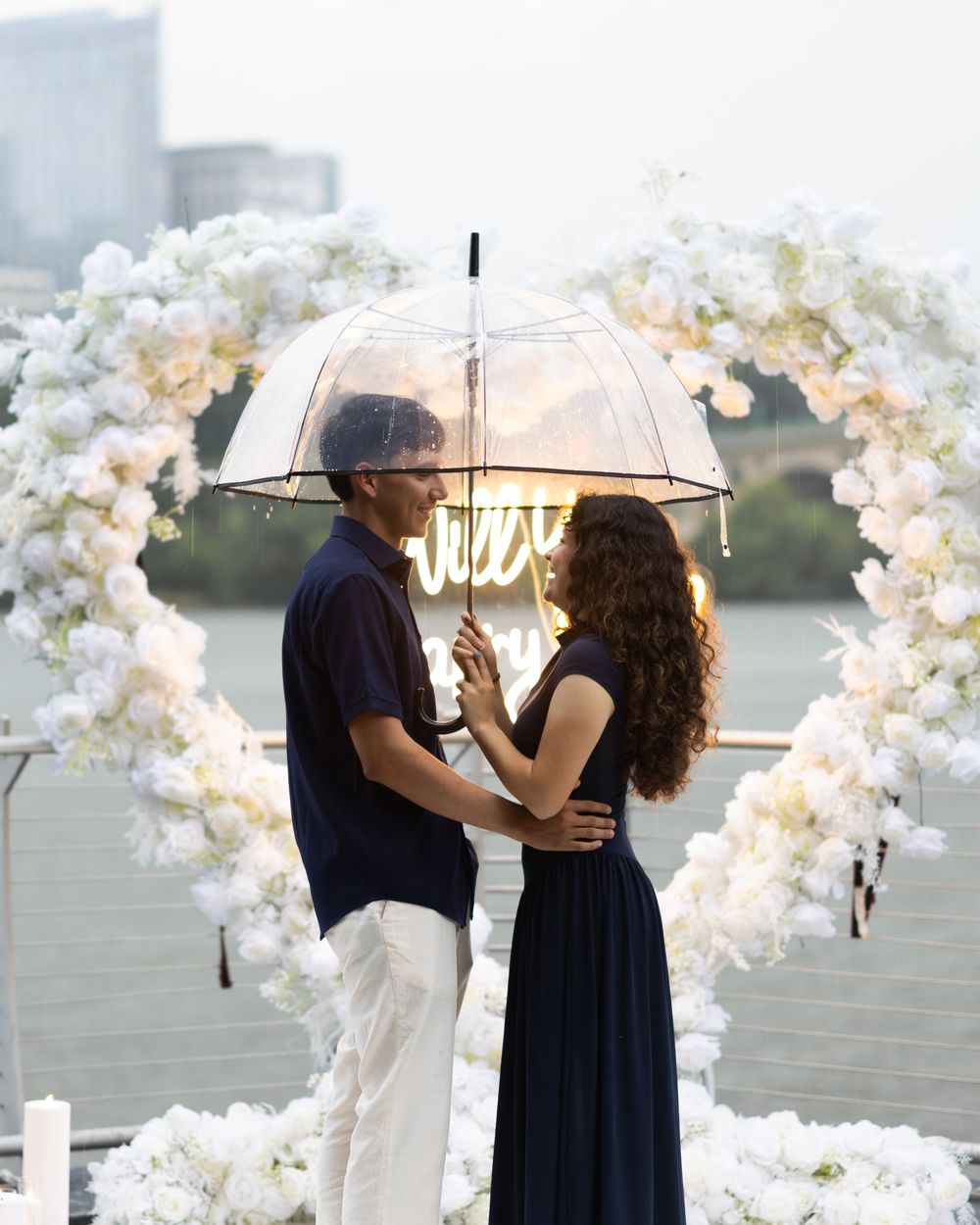 Couple sharing a romantic moment under a clear umbrella framed by a white floral arch near waterfront.