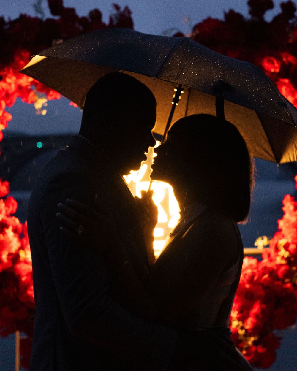 Romantic couple silhouetted under umbrella in rain, surrounded by red floral arch with warm glowing backlight at night.