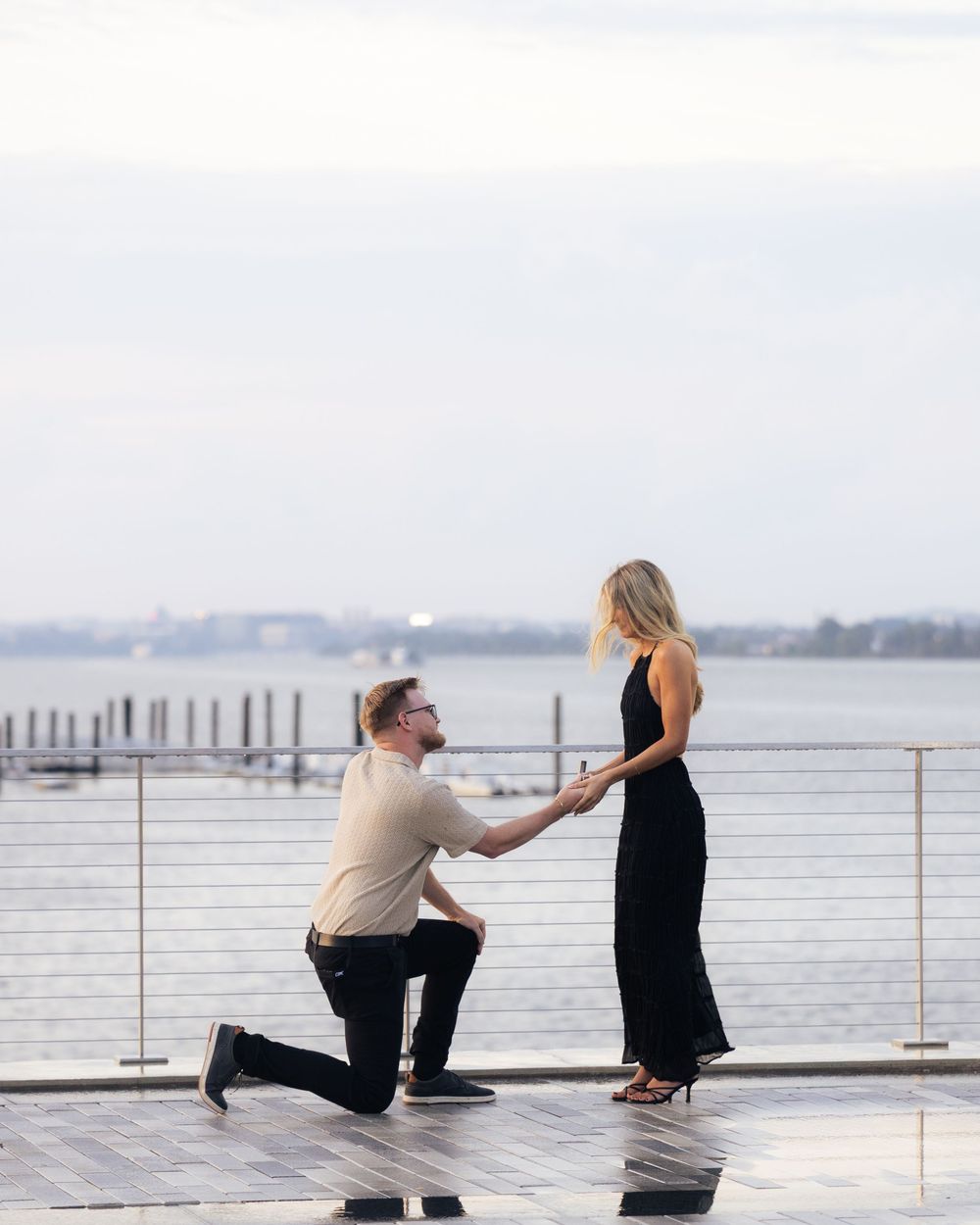 Man proposing on one knee to woman in black dress on waterfront deck at dusk.