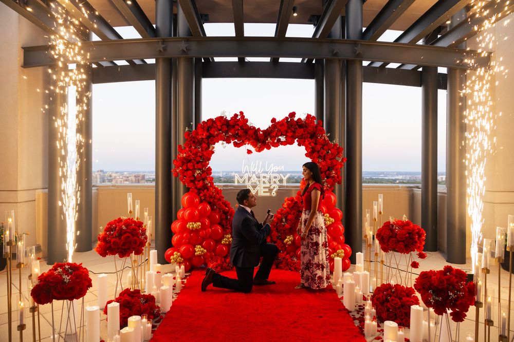 Man proposing on red carpet under heart-shaped floral arch with sparkling lights and red roses at romantic rooftop venue.