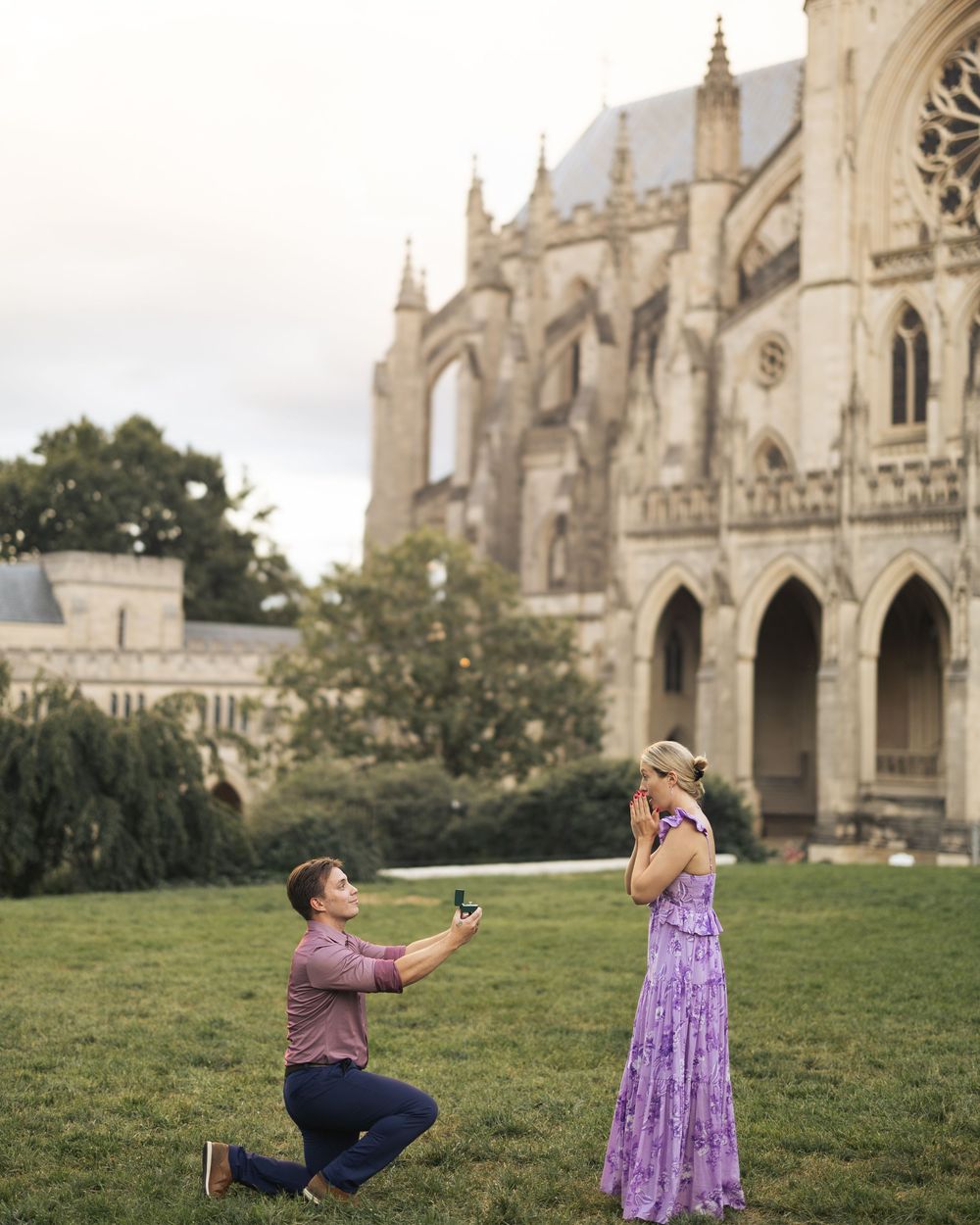 Man proposes to woman on one knee in front of a Gothic cathedral on green lawn, woman covers mouth in surprise.