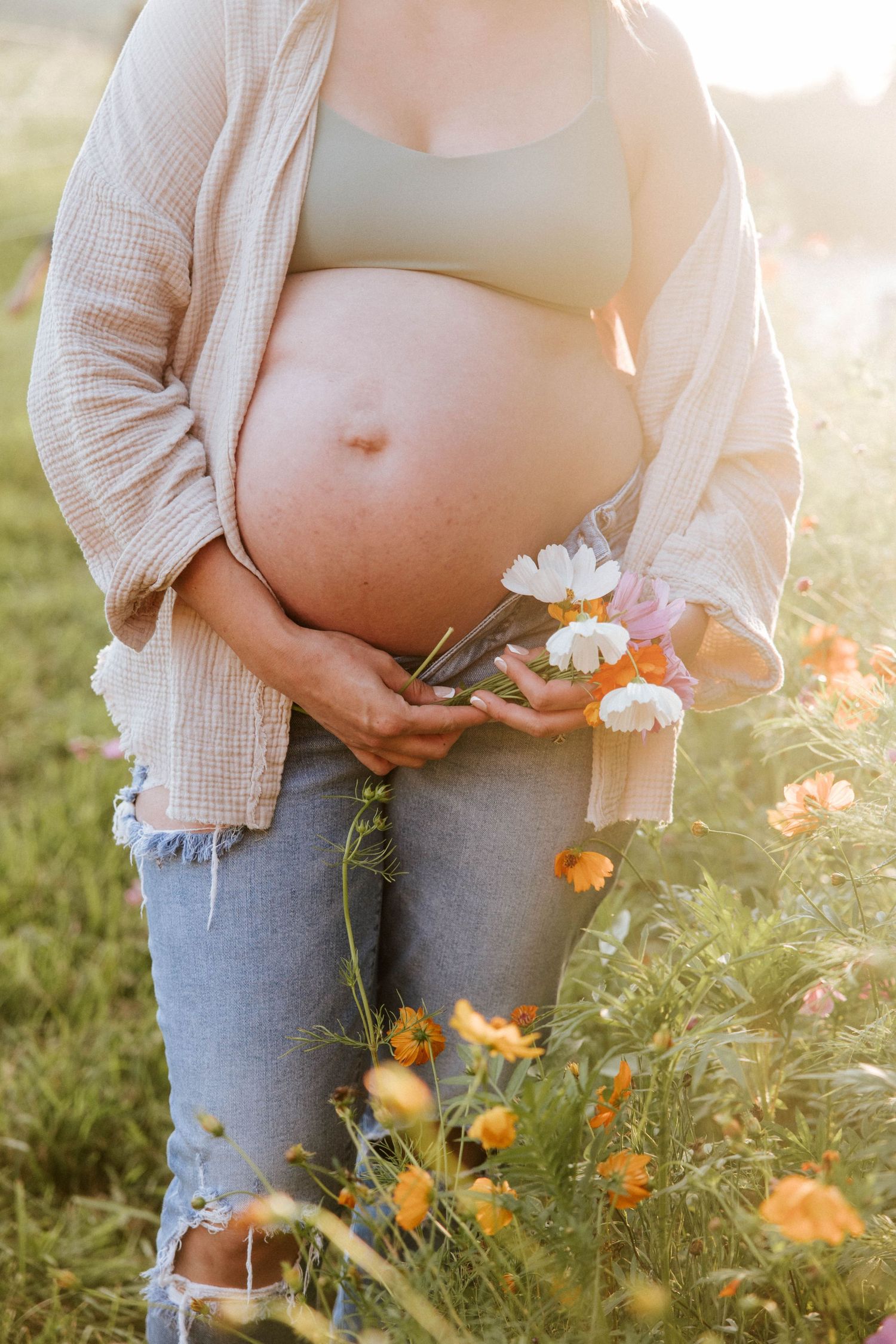 Pregnant woman in ripped jeans holding flowers and ultrasound in sunny field.