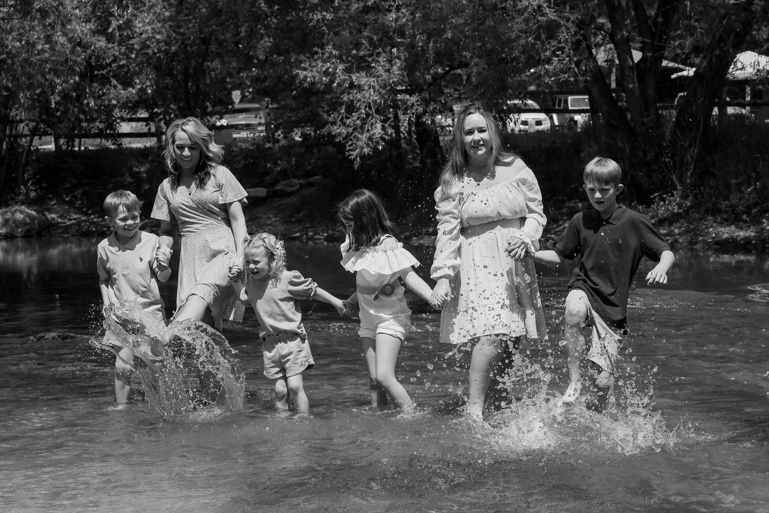 Black and white photo of two women and four children joyfully splashing in a shallow river surrounded by trees.