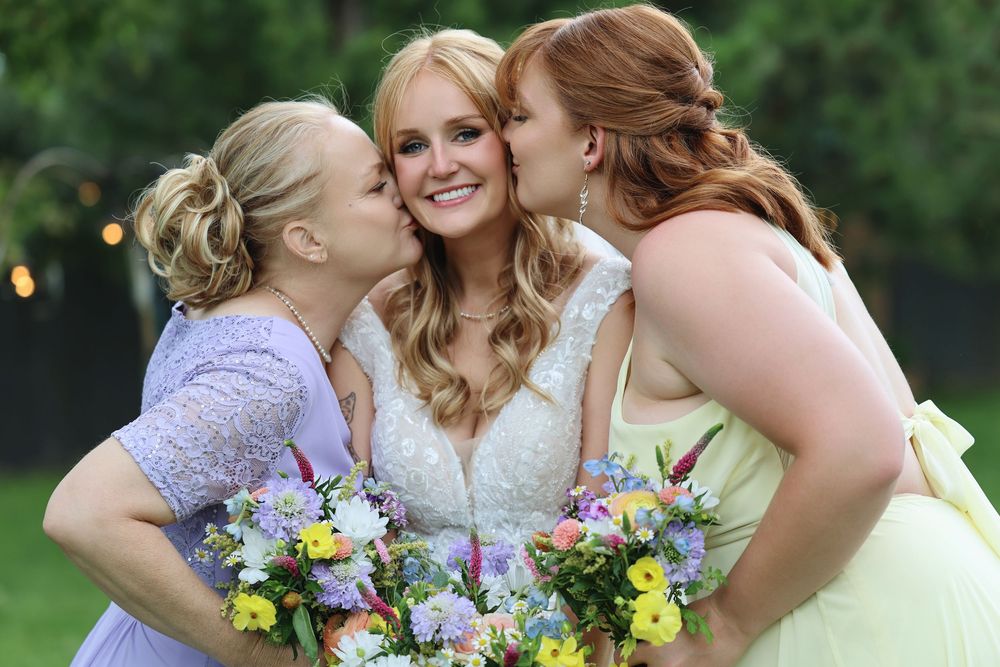 Bride in white lace gown smiles as two bridesmaids in pastel dresses kiss her cheeks, all holding colorful bouquets.