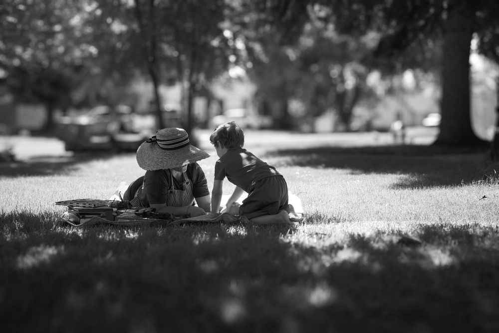 Two children sitting on grass in a park, one wearing a straw hat, sharing a quiet moment in black and white.