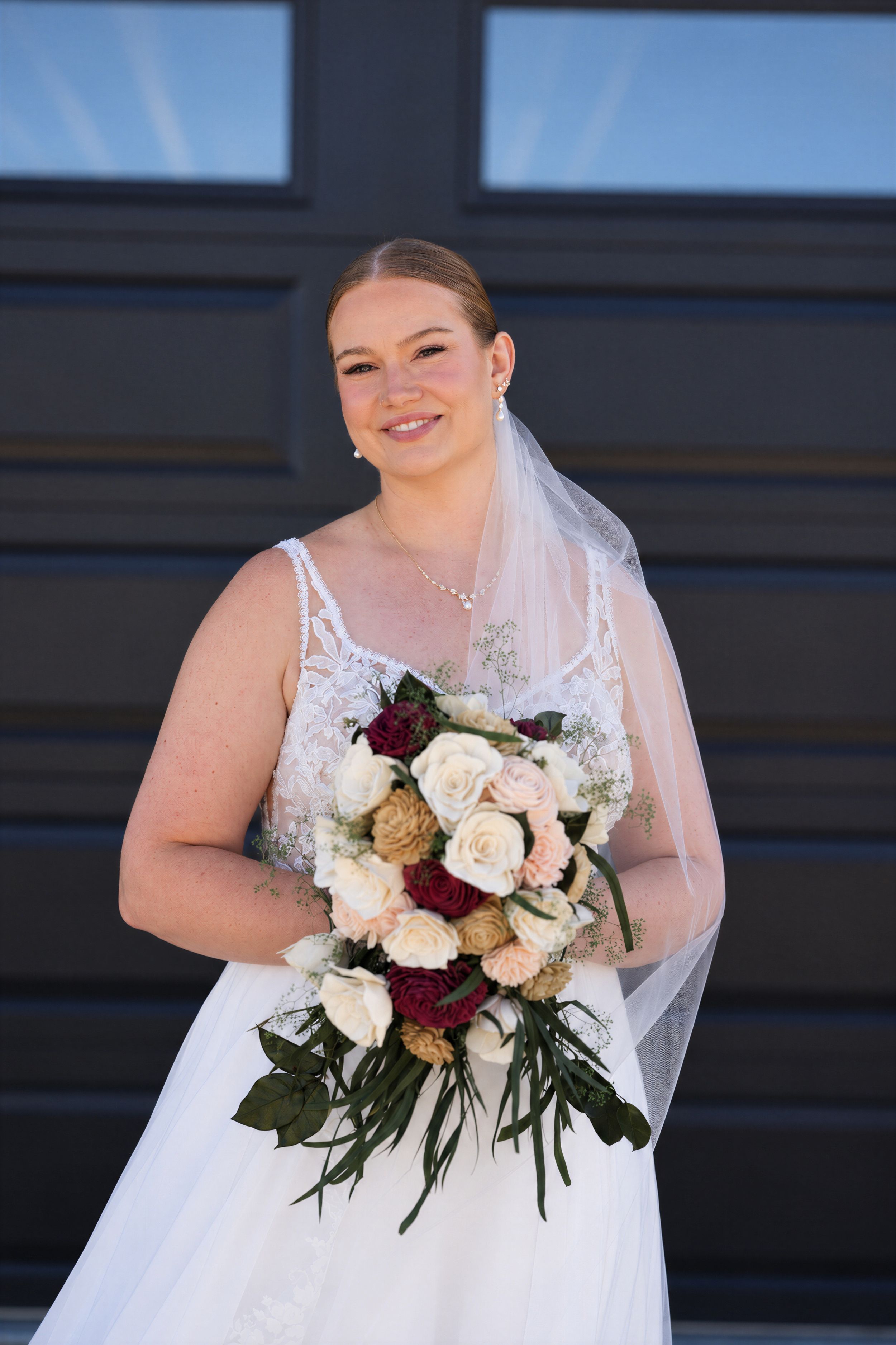 Smiling bride in lace wedding dress and veil holding a bouquet of cream and burgundy roses.