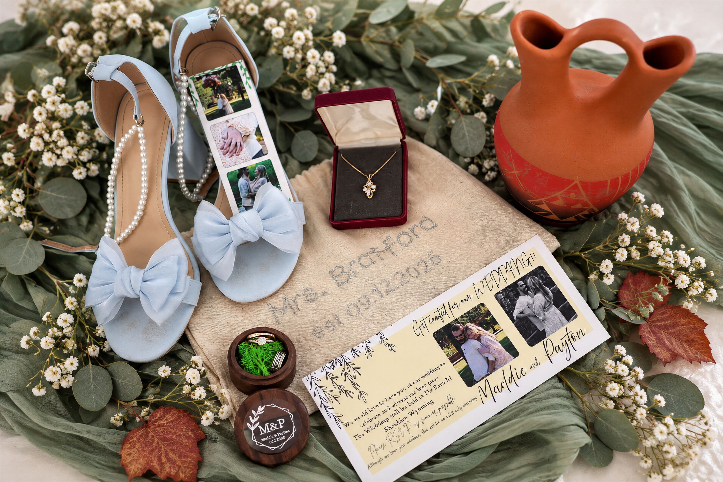 Elegant wedding flat lay with blue bow heels, pearl necklace, gold necklace, invitation, jewelry box, and terracotta vase on greenery.