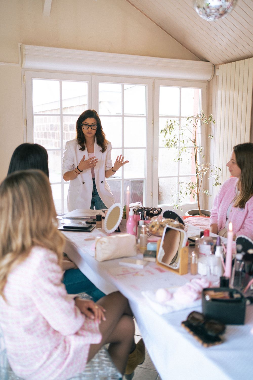Woman in white blazer presenting makeup products to seated attendees at a beauty workshop with cosmetics on table.