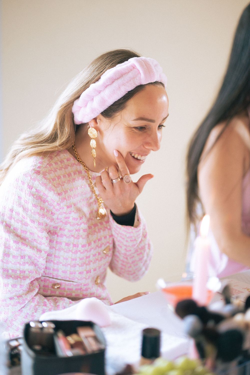 Smiling woman in pink tweed jacket and fluffy headband wearing gold earrings and necklace at a makeup table.