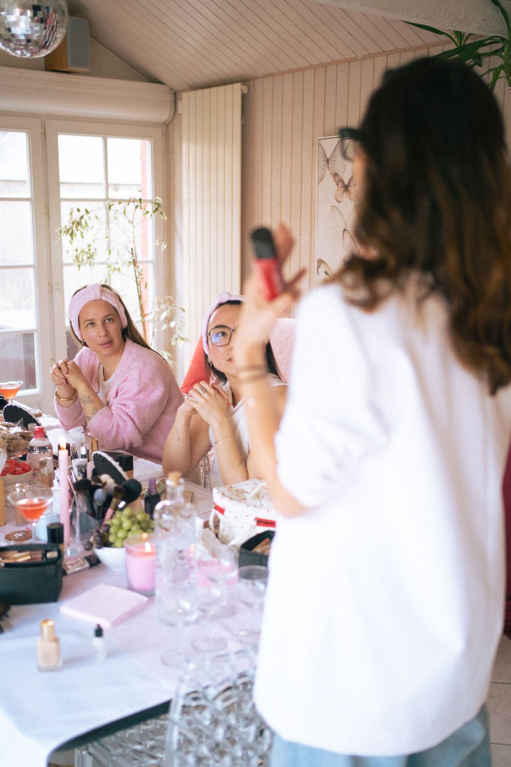 Three women enjoy a beauty spa day, applying makeup at a table filled with cosmetics and glassware indoors.