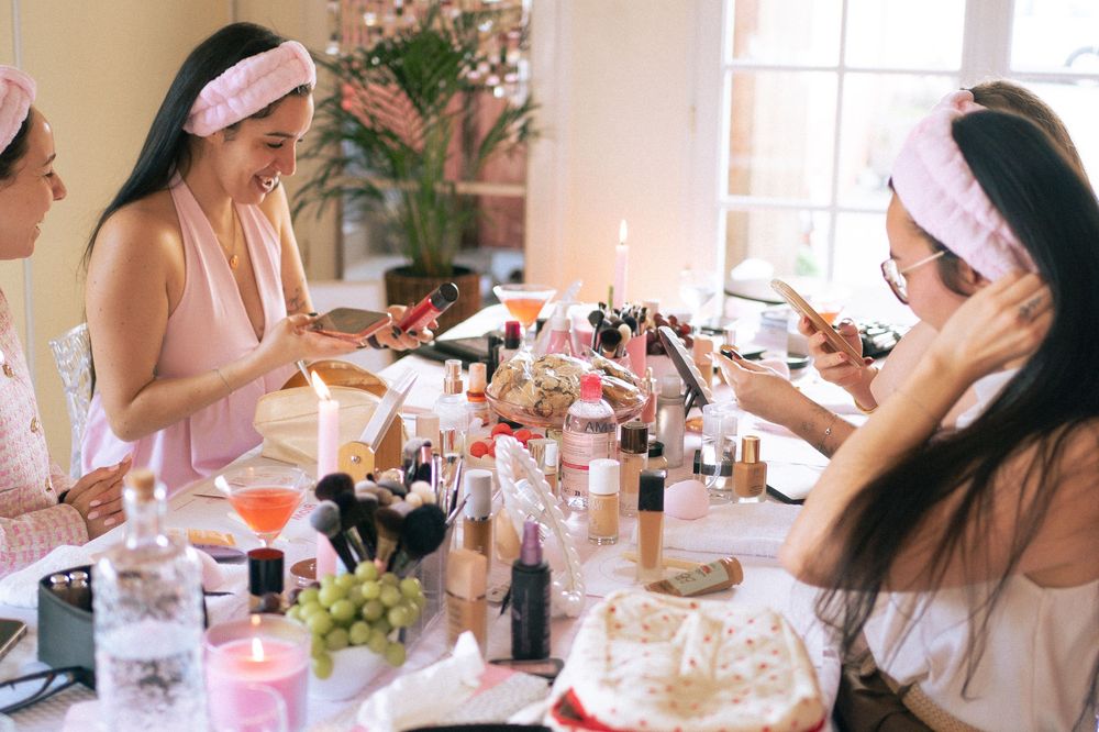 Women wearing pink headbands gather around a table covered in makeup products at a beauty party.