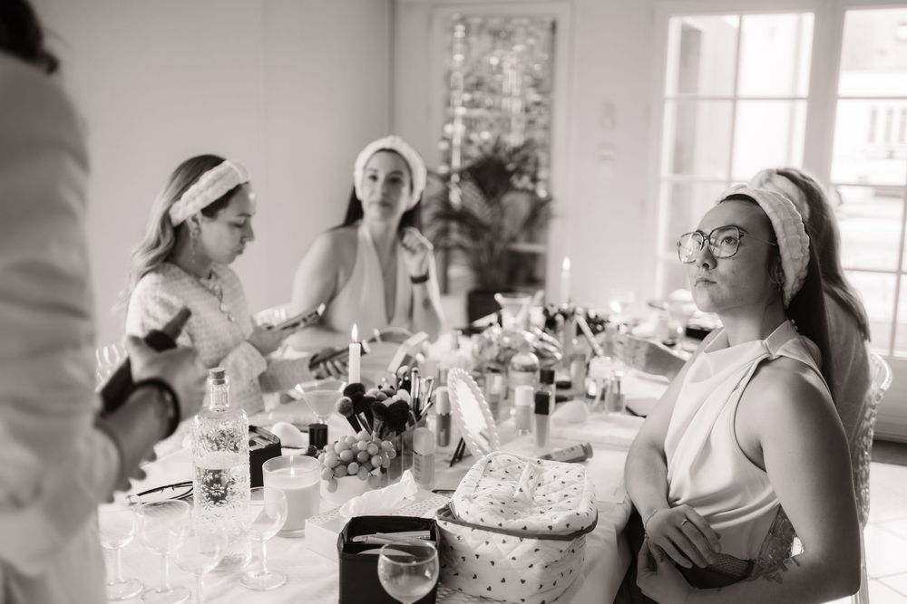 Black and white photo of women with headbands gathered around a table filled with beauty products during a bridal party.