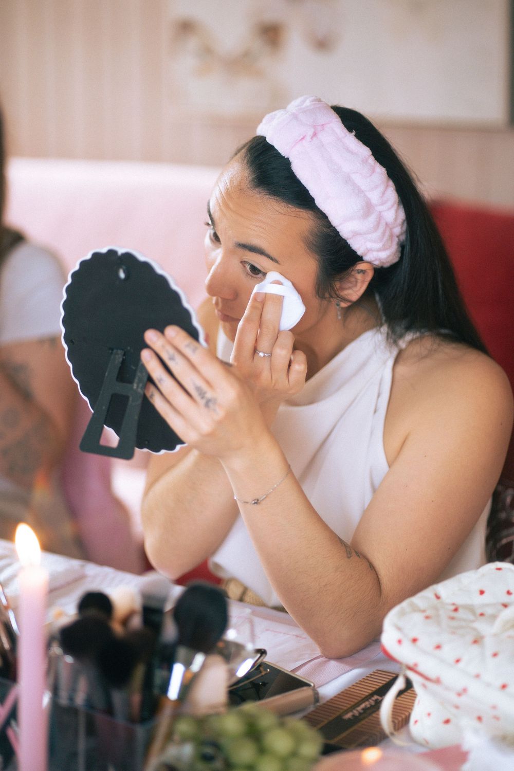 Woman applying makeup remover with cotton pad while holding mirror, wearing pink headband at vanity table.