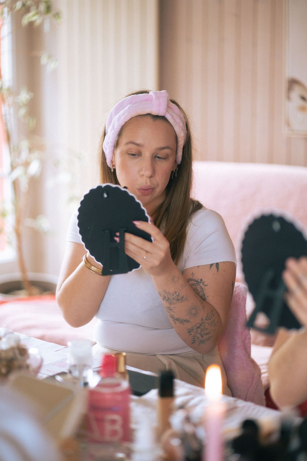 Young tattooed woman with pink headband examining makeup in black mirror at vanity table with beauty products.