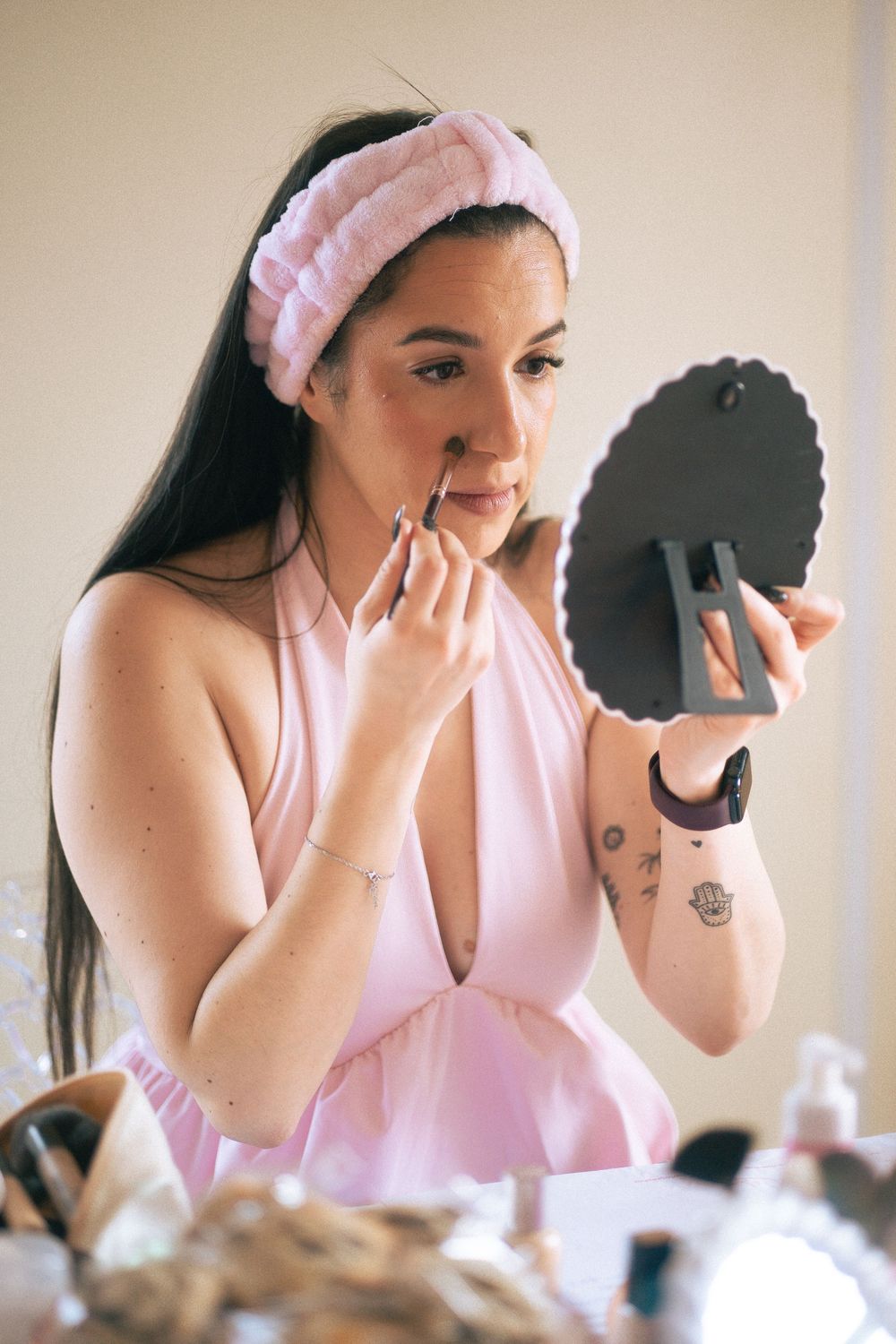 Young woman with pink headband applies eye makeup using a handheld mirror at a beauty vanity table.