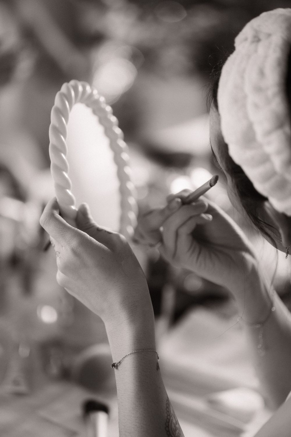 Black and white photo of a woman applying makeup while holding a mirror, wearing a knit beanie hat.