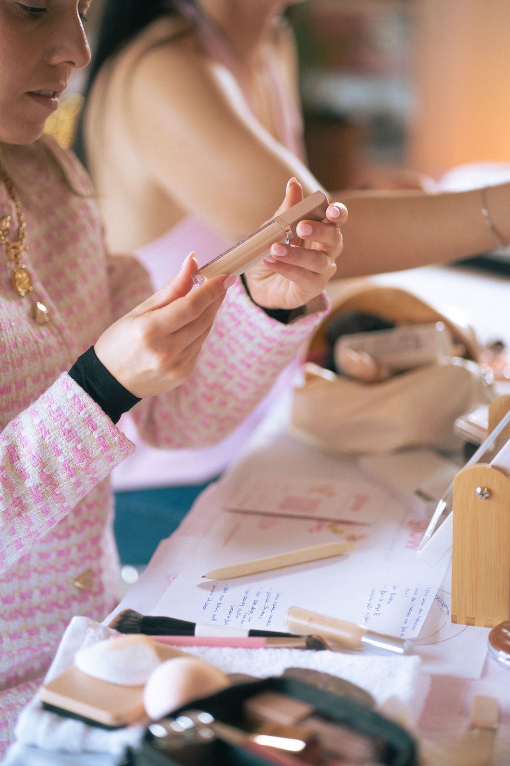 Woman in pink outfit examining makeup brush at beauty table with cosmetics and pencils.