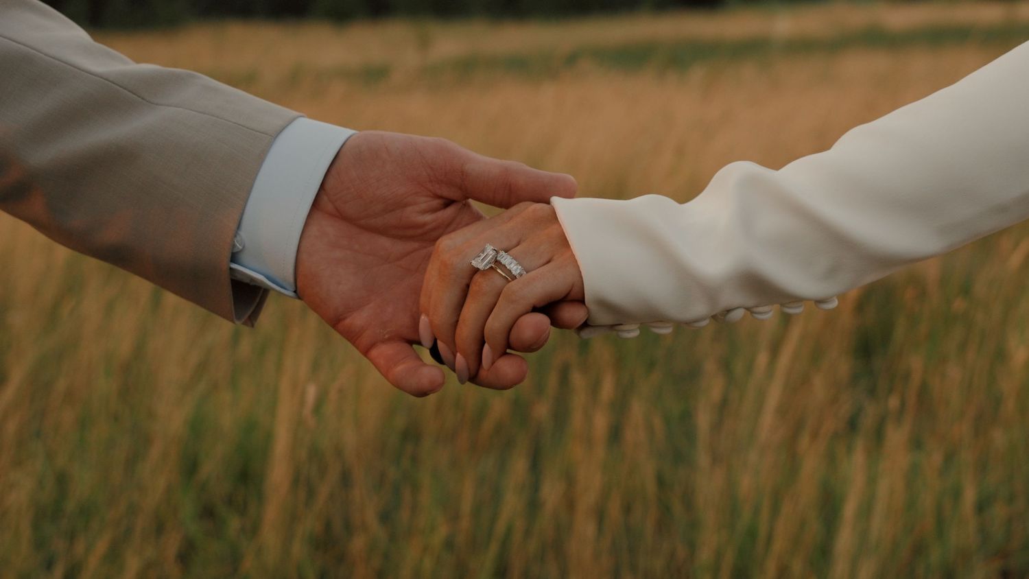 Couple in wedding attire holding hands in a golden field with mountains, bride in white gown and veil, groom in tan suit.
