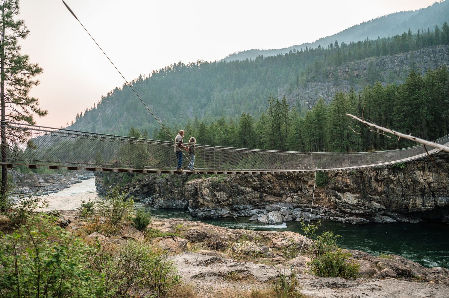 Couple standing on a rustic suspension bridge over a river surrounded by forested mountains.