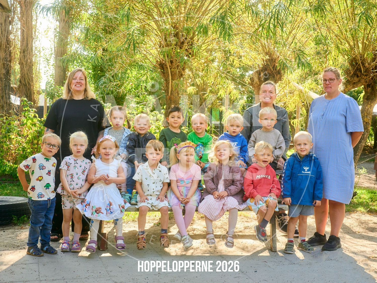 Group photo of children and teachers from Hoppelopperne 2026 posing outdoors among lush green trees.
