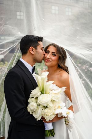 Groom kisses bride on cheek under flowing veil as she smiles holding white roses and calla lily bouquet.