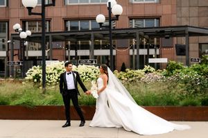 Bride and groom hold hands outdoors in front of modern building, bride in white gown with long train, groom in black tuxedo.