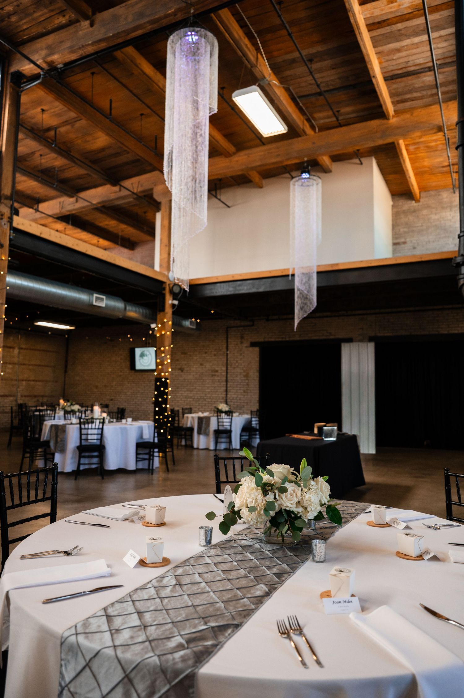 Elegant wedding reception hall with crystal chandeliers, white round tables, floral centerpieces, and warm wood ceiling accents.