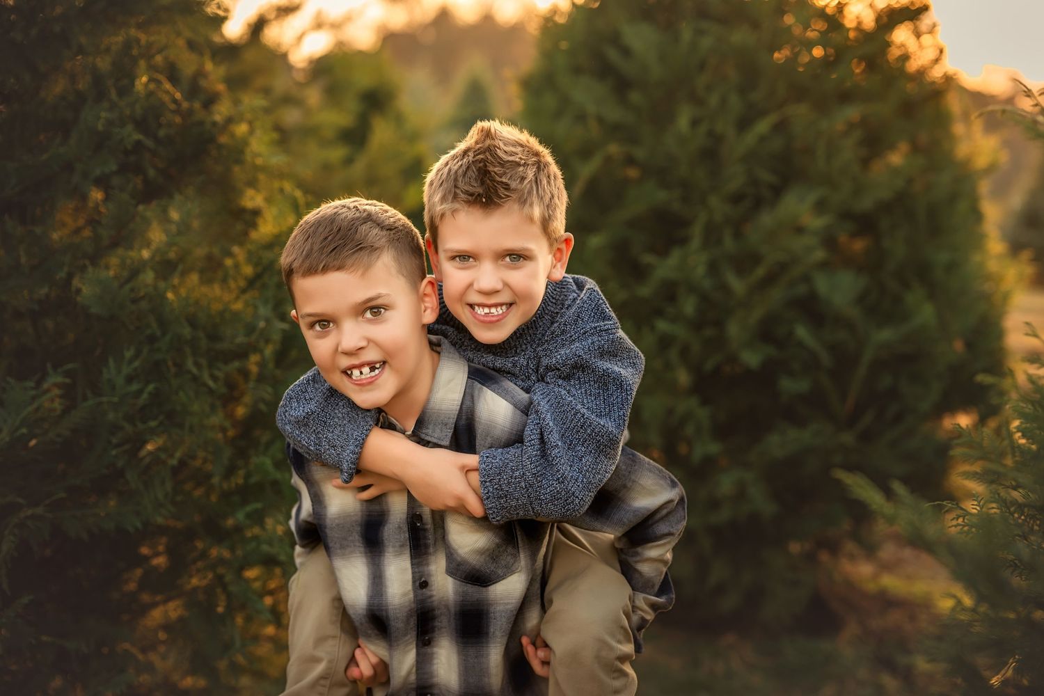 Two young boys pose for a photo at a Christmas tree farm, one giving the other a piggyback ride at sunset.