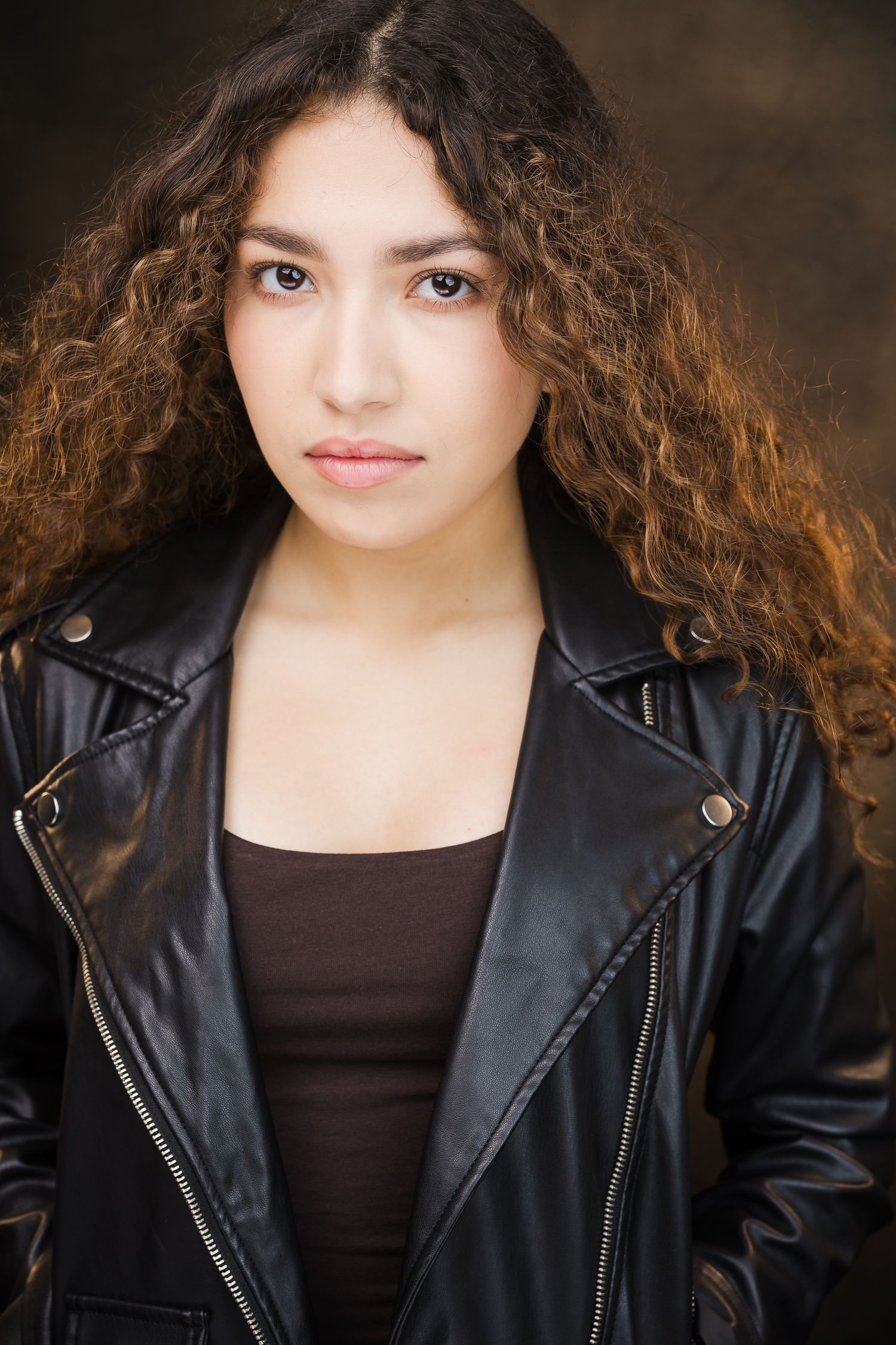 Woman in black leather jacket with arms crossed, posing confidently against a gray background.