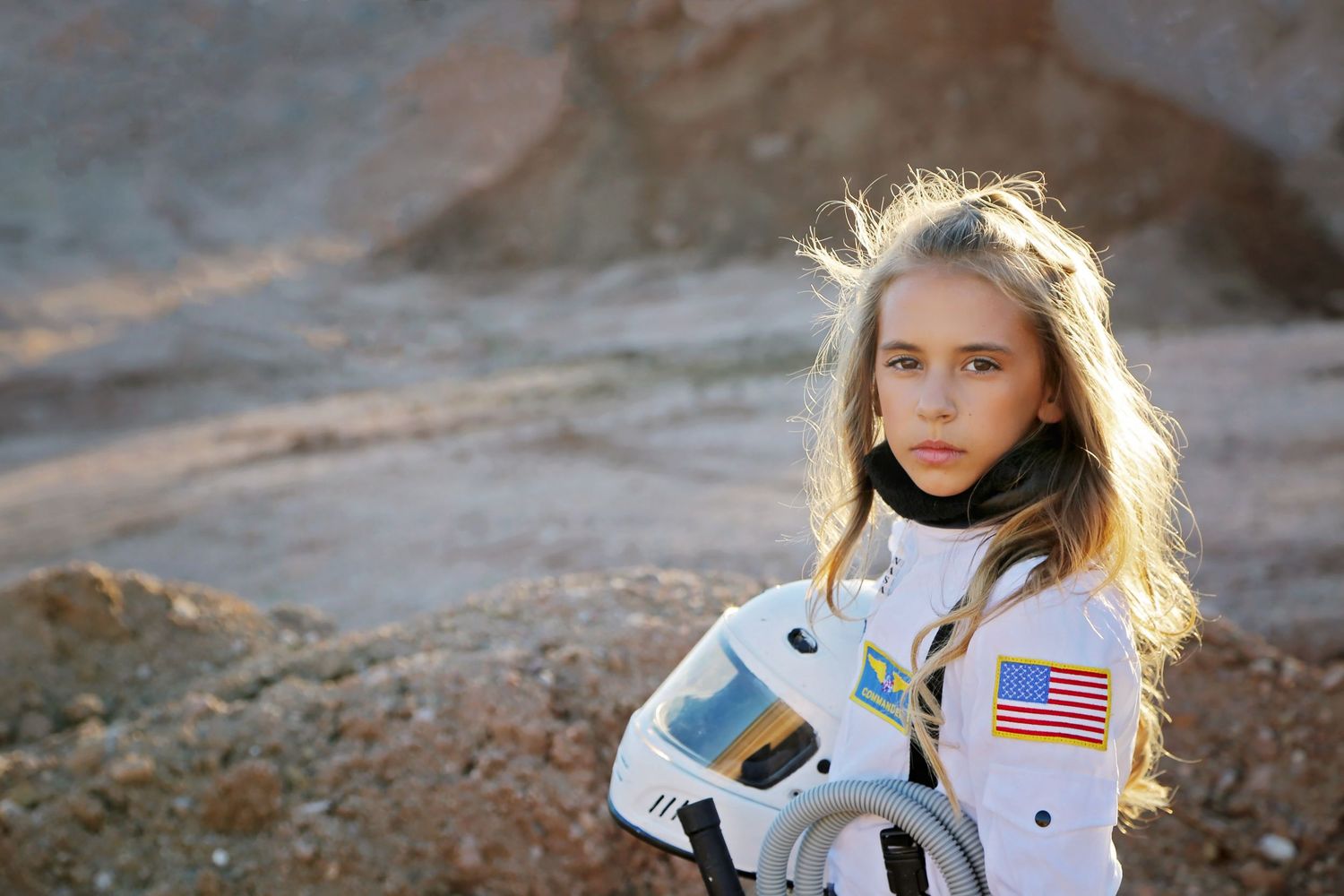 A young girl with long blonde hair wears a white astronaut suit with an American flag patch in a rocky Mars-like landscape.