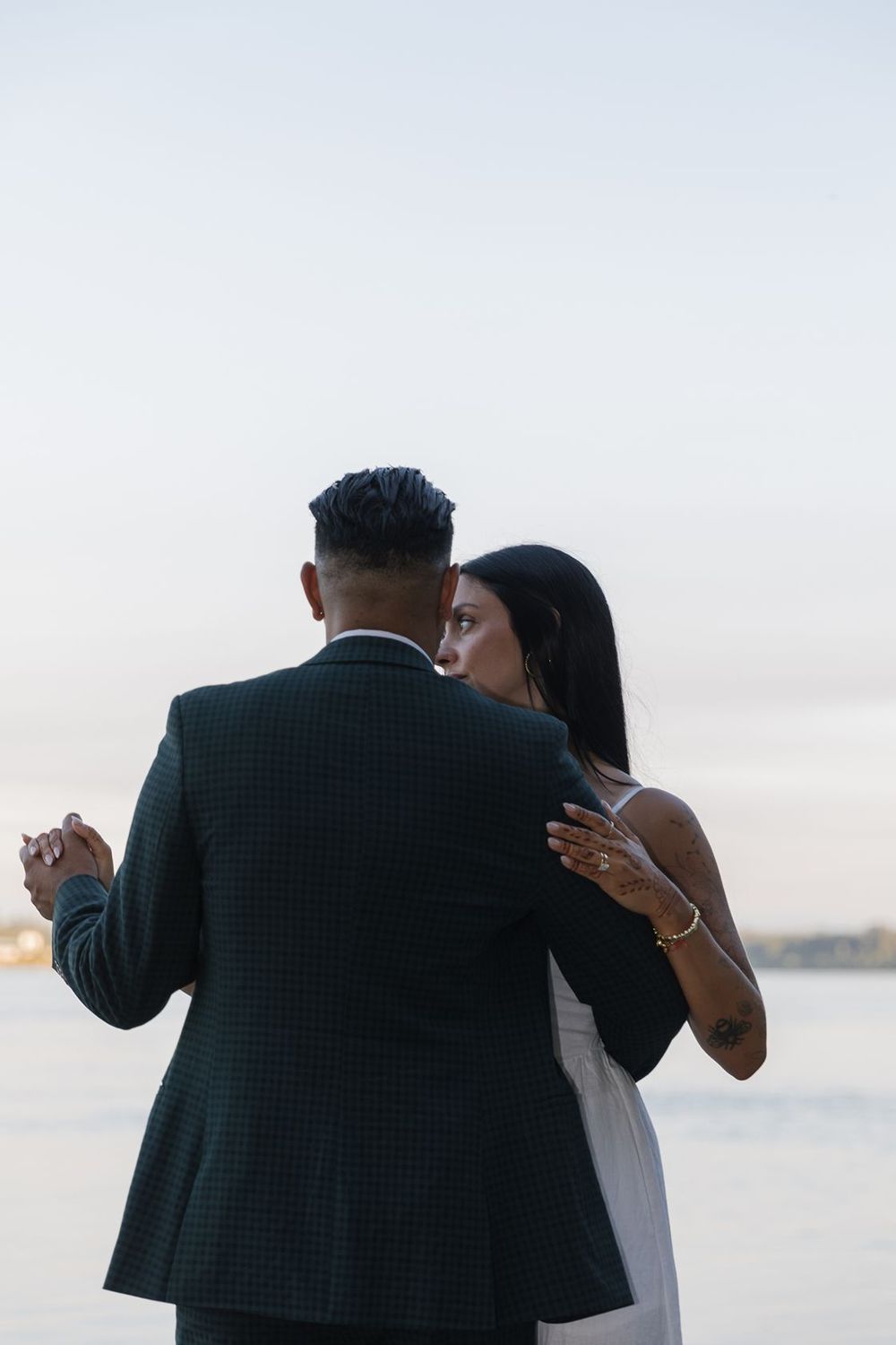 Couple dancing outdoors near water at dusk, woman in white dress embracing man in dark suit.