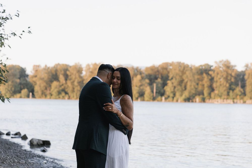 Couple embracing by a lakeside at dusk, man in dark suit, woman in white dress, with autumn trees in background.
