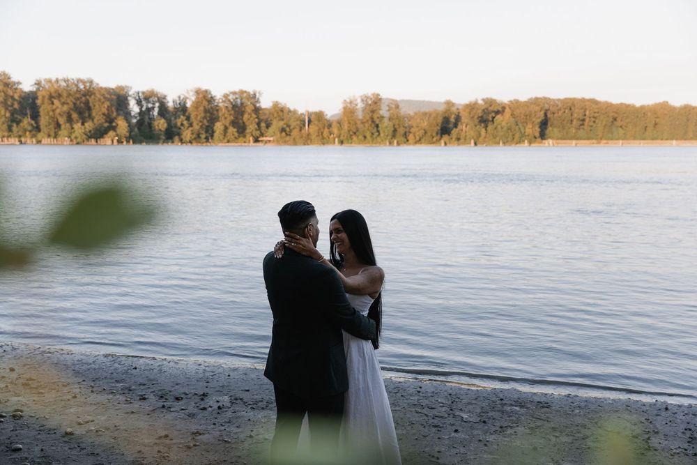 Couple embracing in wedding attire on a lakeside beach with trees and calm water in the background.