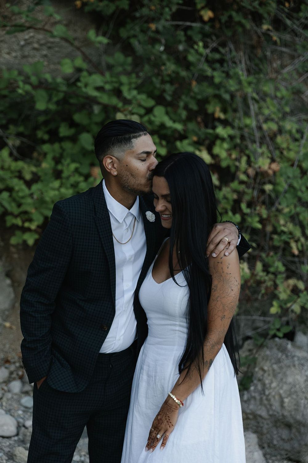 Groom in dark suit kisses bride's forehead as she smiles in white dress outdoors.