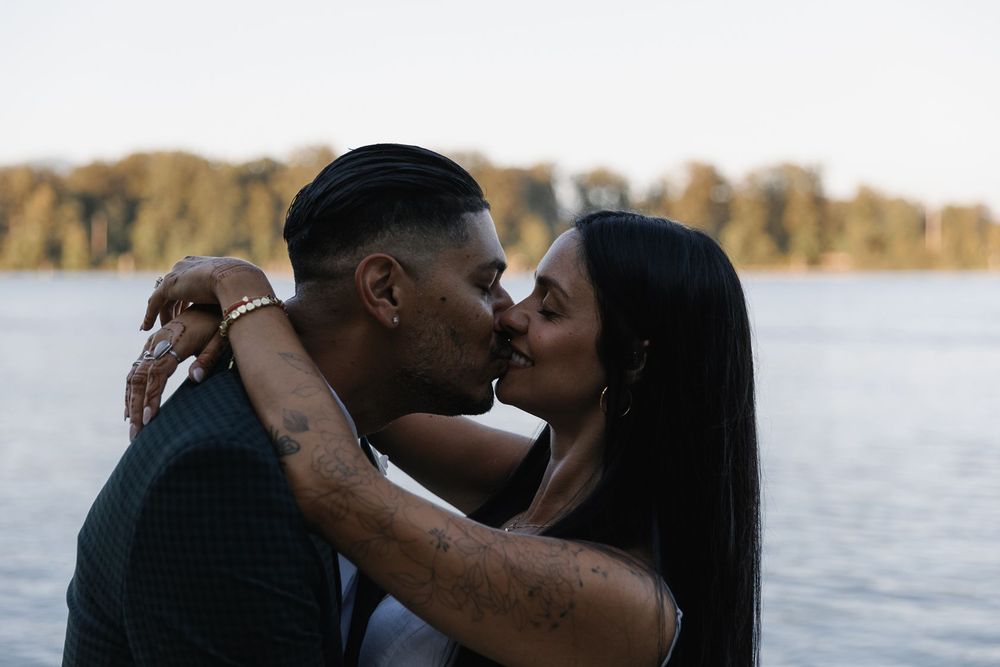 A couple sharing a romantic kiss by a lakeside with autumn trees in the background.