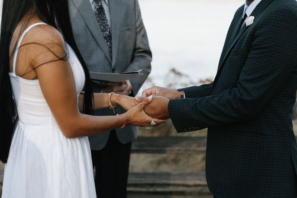 Couple exchanging vows and holding hands during an outdoor wedding ceremony with an officiant in background.